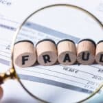 Magnifying glass over a financial document with 'FRAUD' spelled out in wooden blocks.