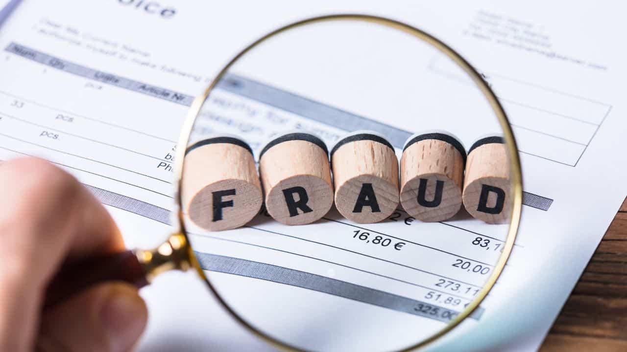 Magnifying glass over a financial document with 'FRAUD' spelled out in wooden blocks.