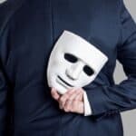 Business person in a suit holding a white mask behind their back.