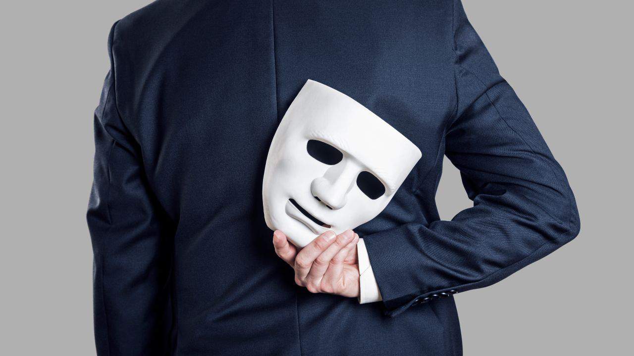 Business person in a suit holding a white mask behind their back.