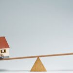 Miniature house and stacked coins on a seesaw representing property investment.