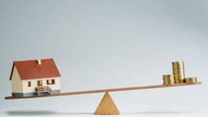 Miniature house and stacked coins on a seesaw representing property investment.