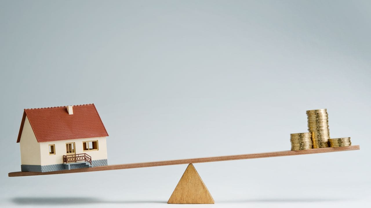 Miniature house and stacked coins on a seesaw representing property investment.