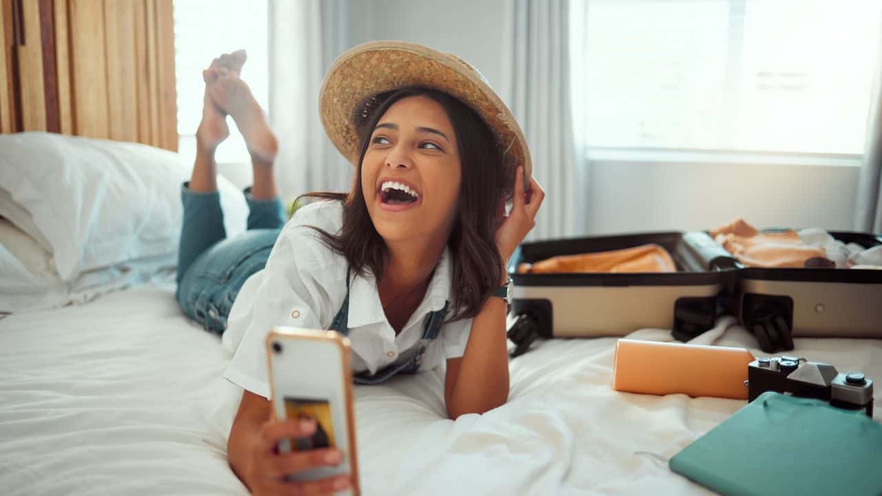 Smiling woman with a hat using her phone in a hotel room.