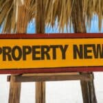 Property news sign on beach with blue sky and ocean background.