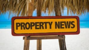 Property news sign on beach with blue sky and ocean background.