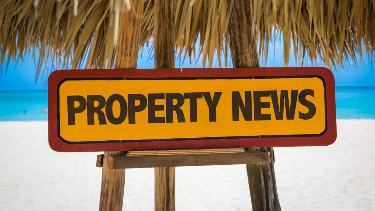 Property news sign on beach with blue sky and ocean background.